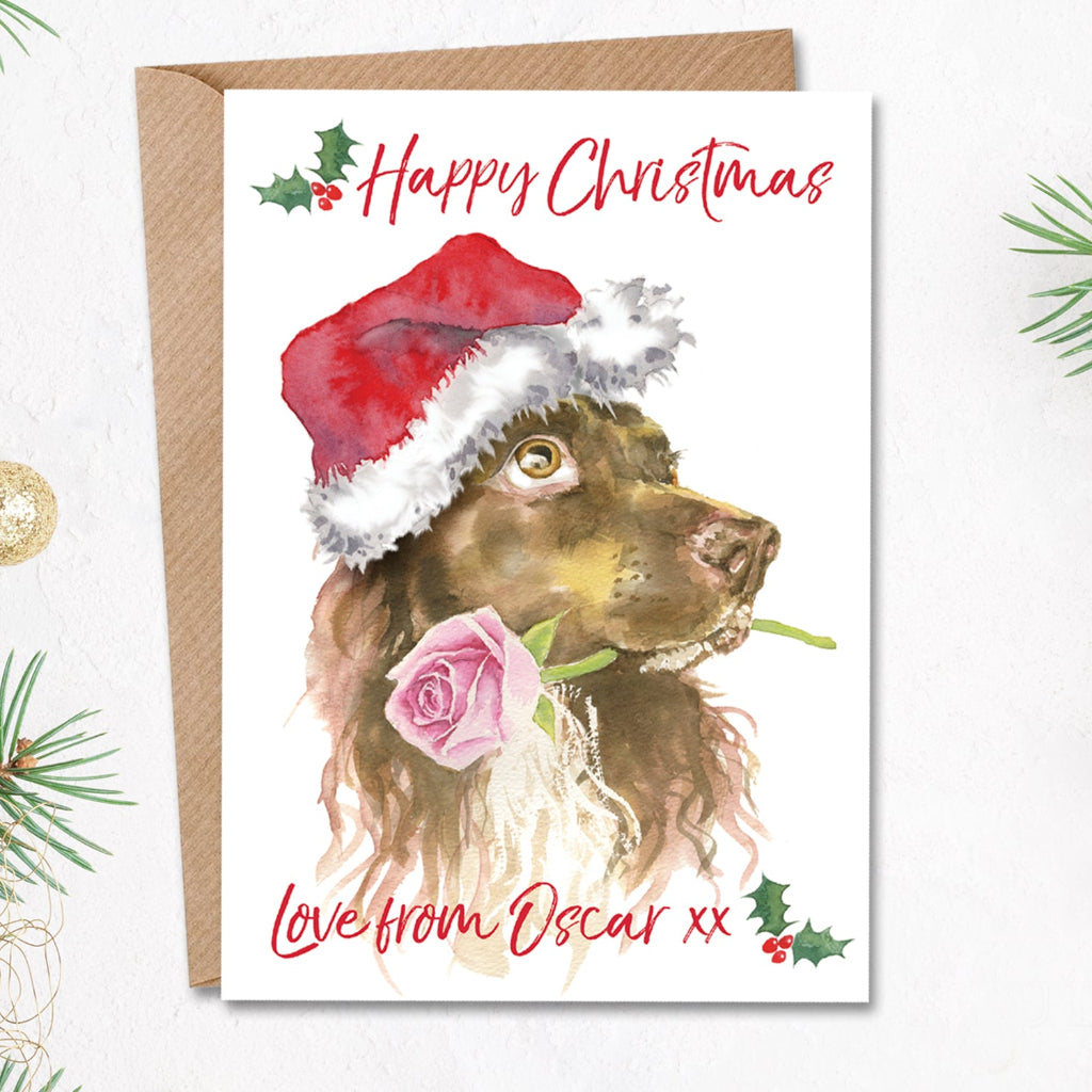 Personalised christmas card with a liver brown working cocker spaniel in a santa hat holding a pink rose in his mouth on the front. The cute xmas card has the dog's name printed on the front