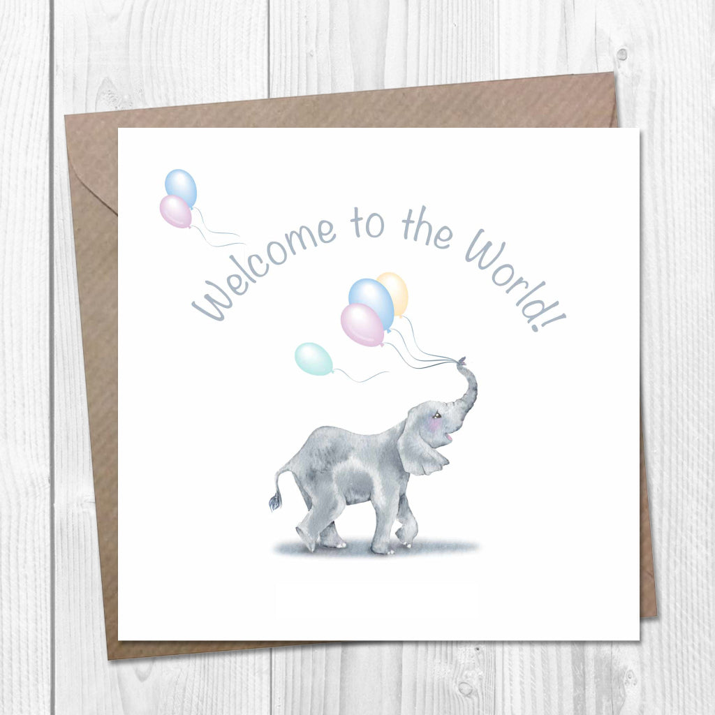 A cute baby elephant holds pastel balloons with its trunk, under the words "Welcome to the World!" on a card set against a wooden background.