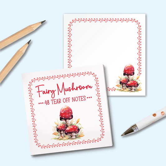 A set of square tear-off notes with a red border and a watercolor illustration of red mushrooms, shown on a light blue background with two pencils and a white pen as props.