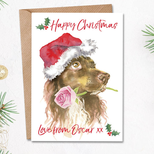Personalised christmas card with a liver brown working cocker spaniel in a santa hat holding a pink rose in his mouth on the front. The cute xmas card has the dog's name printed on the front