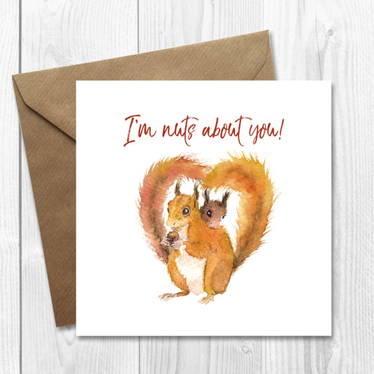 A Valentine's Day or Love you card featuring a watercolor illustration of two red squirrels forming a heart shape with their tails, and the text 'I'm nuts about you!'. The card is placed on a wooden surface with a brown kraft envelope.