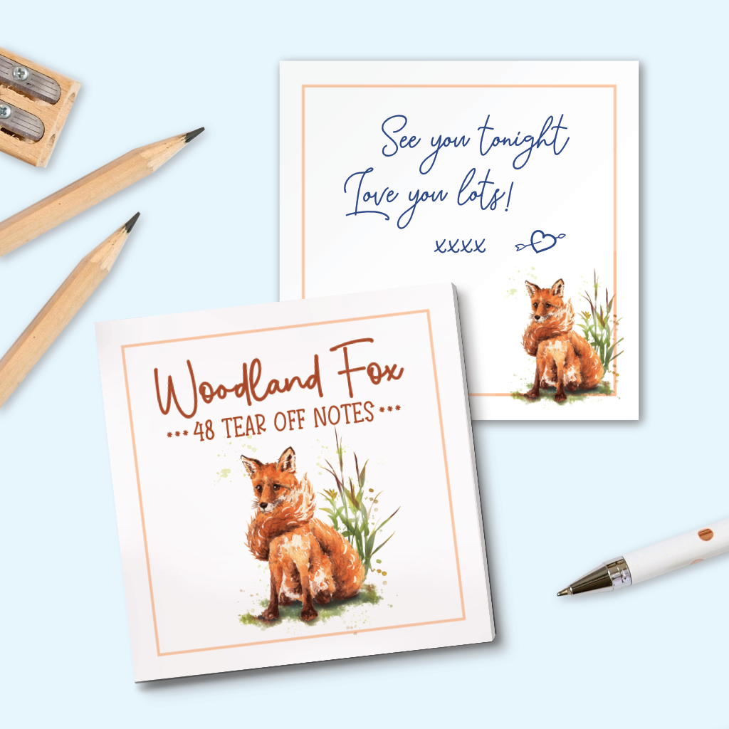 Two square tear-off notes with whimsical fox illustrations are arranged on a light blue background with pencils and a pen as props. One page has a love note written on it.